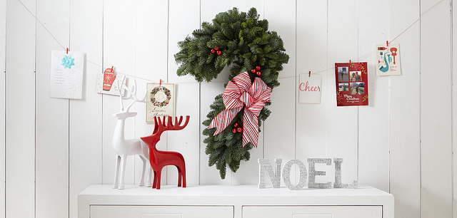 Pine Christmas wreath shaped like a candy cane with a striped bow over reindeer decorations a noel sign on a table and Christmas cards hanging on a string
